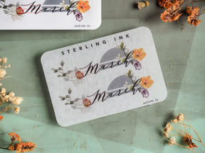 Pale gray planner sticker featuring the word March in delicate script, adorned with vibrant watercolor florals in soft shades of pink, orange, and purple.