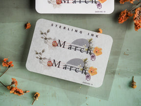 Decorative "March" month sticker featuring delicate watercolor botanical illustrations in soft pastel shades of yellow, pink, and gray.
