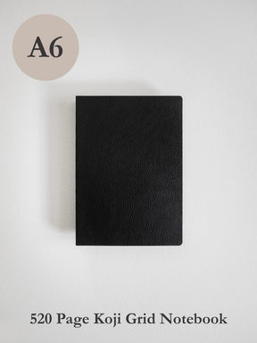 A black textured notebook shown from above on a light gray background, with a beige circle labeled 'A6' in the top left and text at the bottom reading '520 Page Koji Grid Notebook'.
#Color_Onyx
