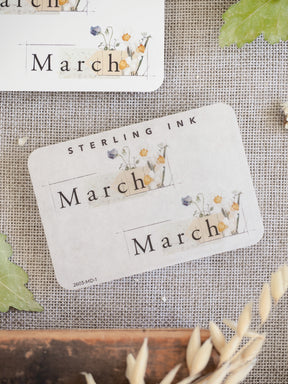 Spring-themed journaling stickers featuring the word "March" surrounded by delicate watercolor depictions of pale florals, buds, and natural greenery.