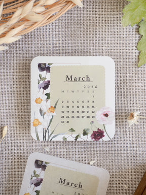 Delicate monthly calendar for March 2026, featuring a soft watercolor floral wreath of muted purples, peaches, and greens surrounding the date grid.