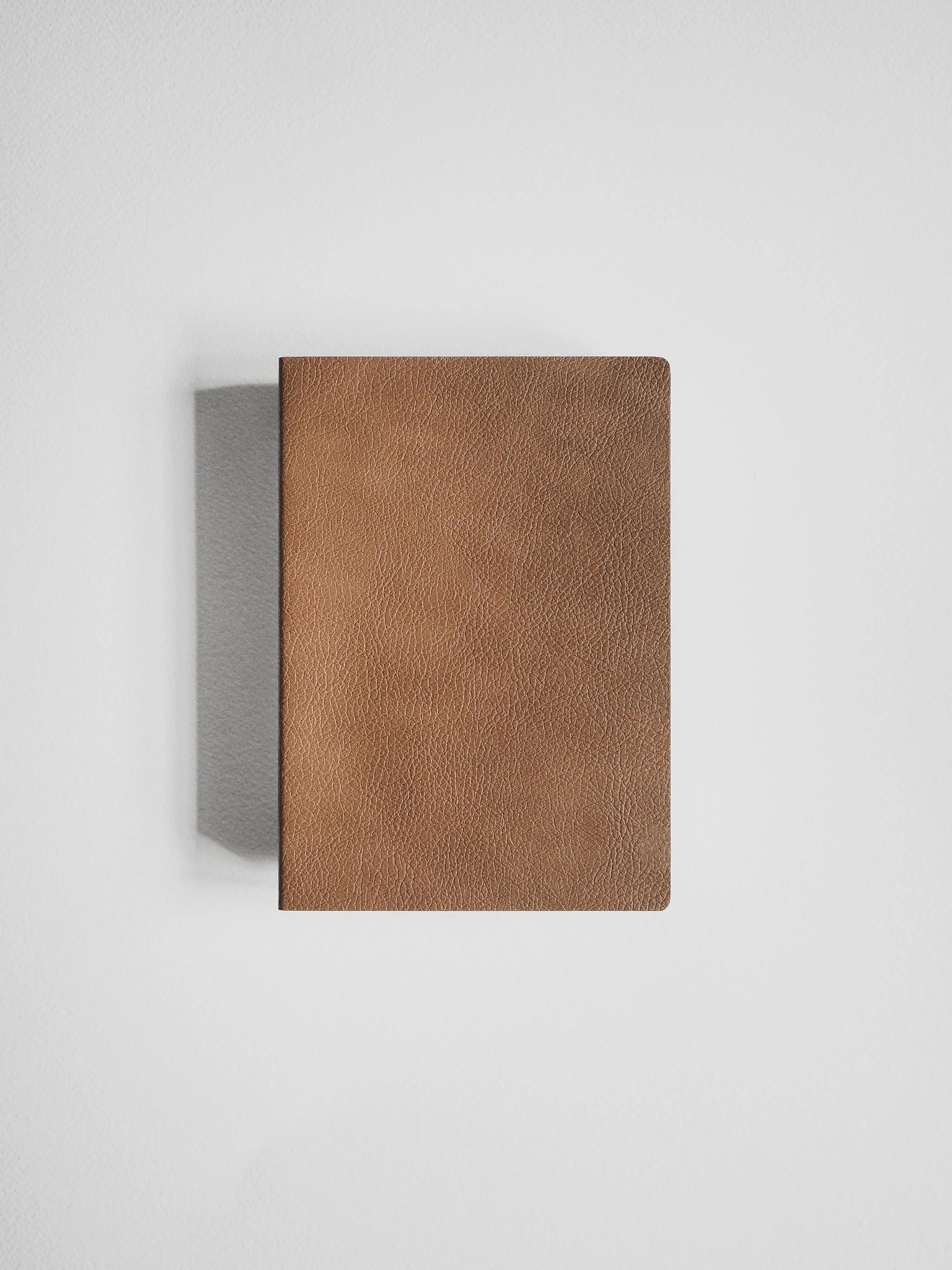 A brown textured leather notebook or journal with rounded corners, shown closed against a light gray background with a soft shadow to the left.
#Color_Caramel
