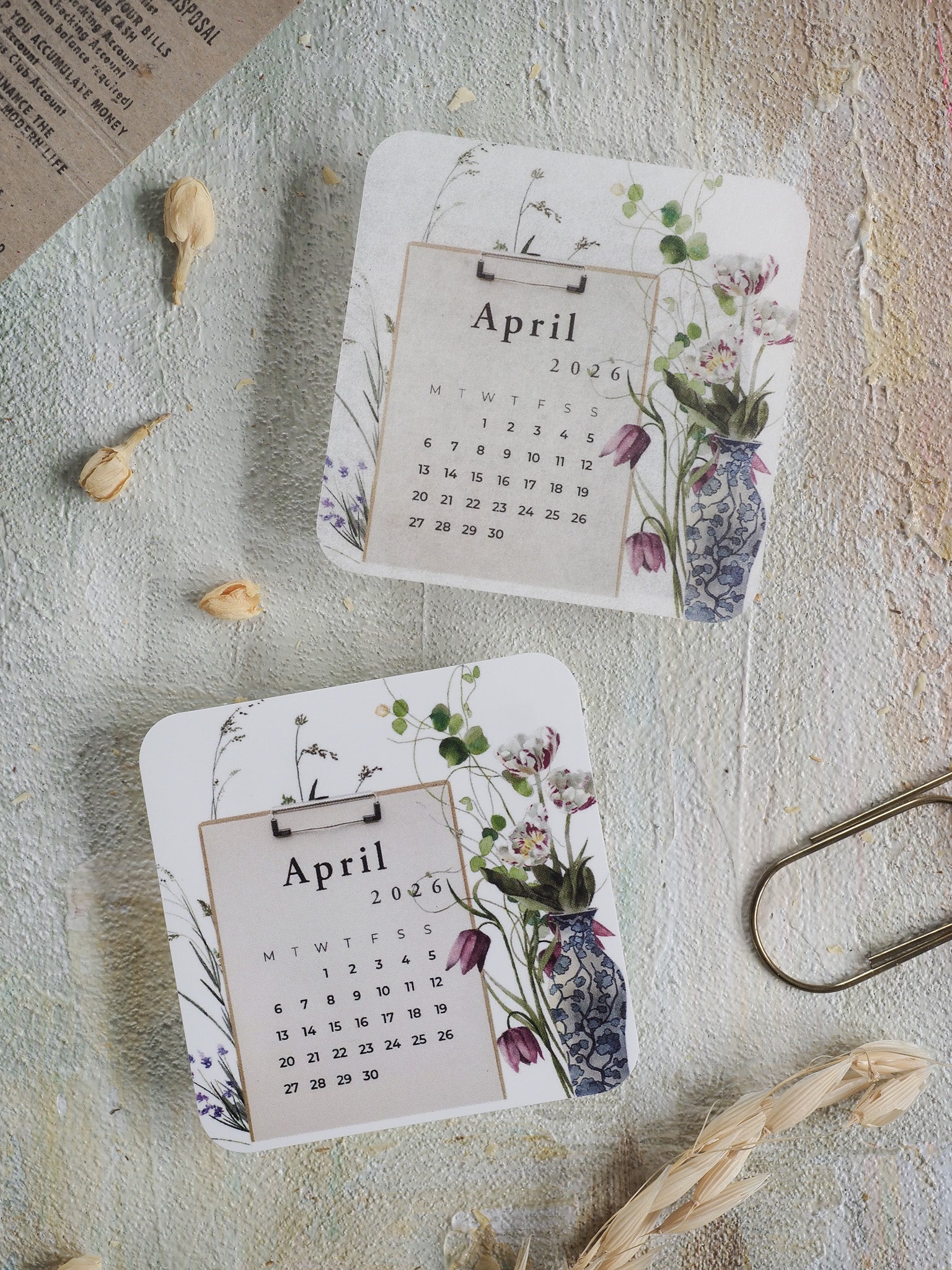 Decorative April 2026 monthly calendar featuring a white background and an elegant botanical wreath border of muted purples, greens, and soft floral illustrations.