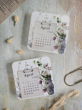 Decorative April 2026 monthly calendar featuring a white background and an elegant botanical wreath border of muted purples, greens, and soft floral illustrations.