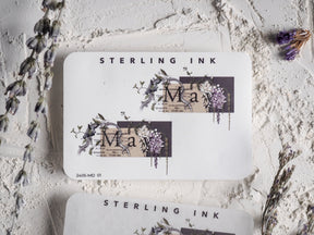 Elegant spring-themed sticker featuring the word "May" surrounded by delicate watercolor clusters of purple, green, and cream floral blooms.