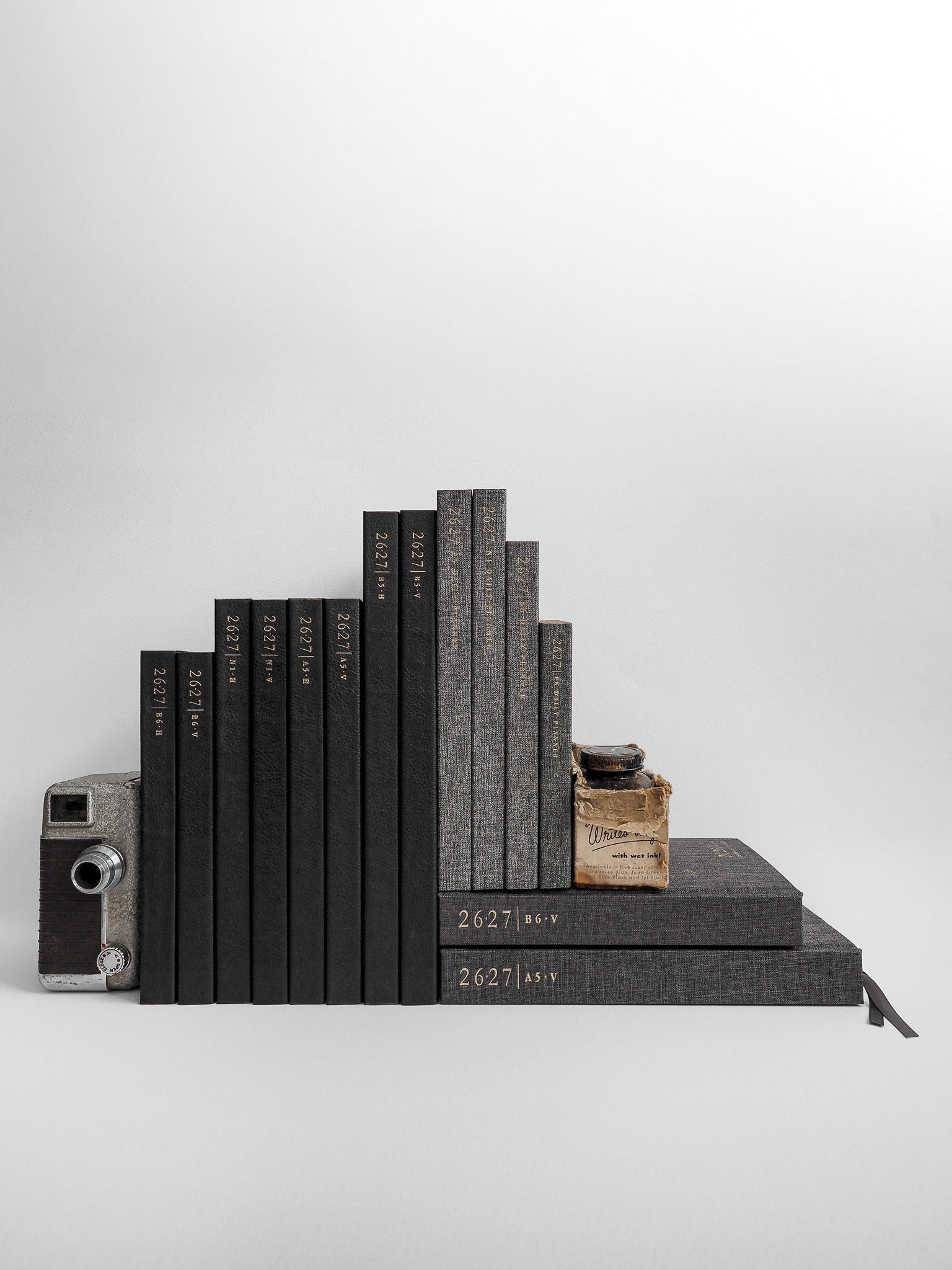 A curated stack of dark gray journals and notebooks, accented by a vintage film camera and small apothecary-style jar.