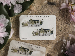 Pale sticker featuring the word "June" framed by a delicate watercolor botanical wreath of green leaves, white, and dusky floral blooms.
