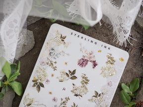 Detailed botanical washi stickers on white paper, featuring clustered designs of soft pink, ivory, and sage green blossoms and leaves with delicate gold foil accents.