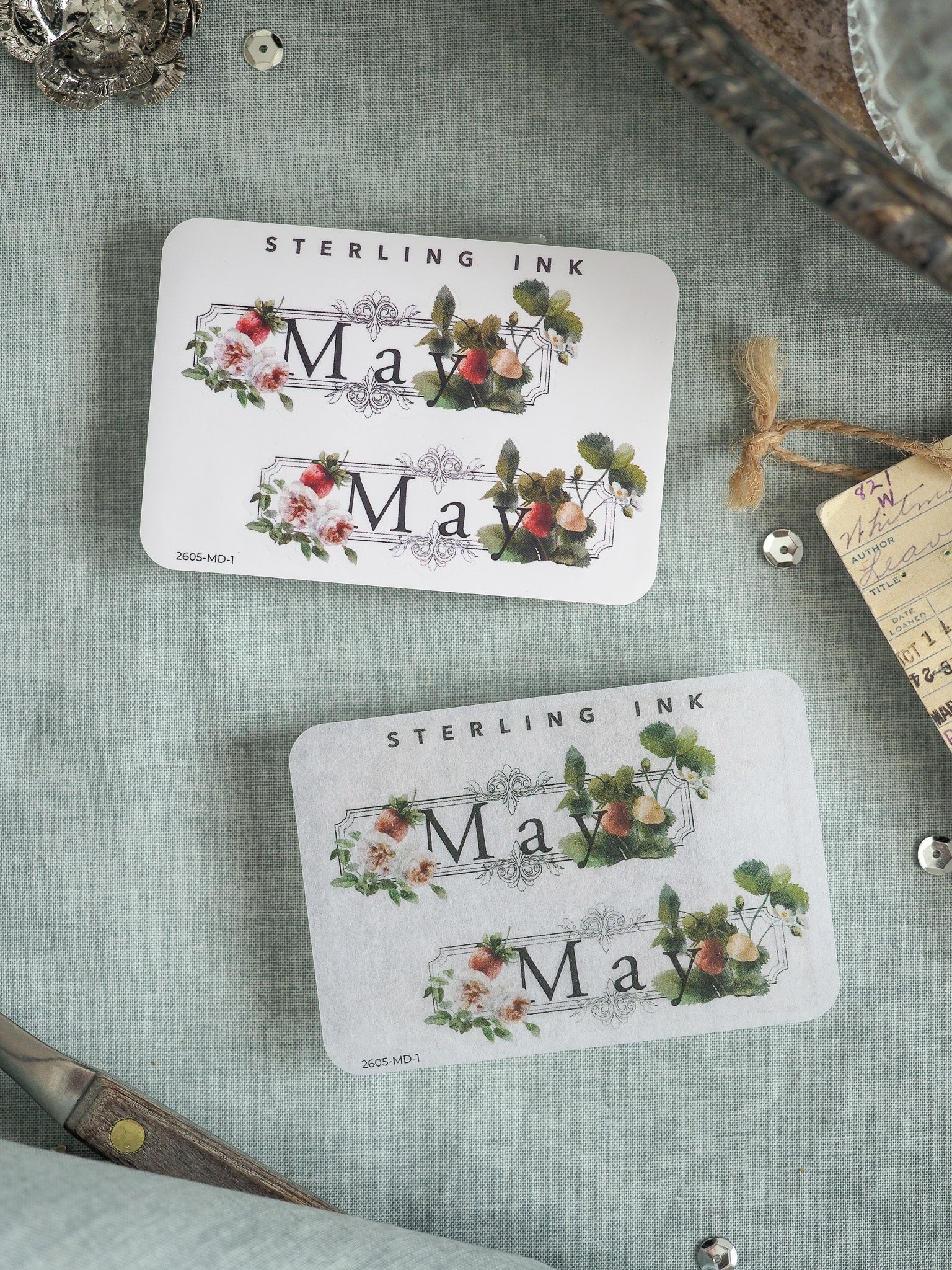 Two delicate, square stickers featuring "May" printed below floral wreaths of pink and red blooms and green foliage, presented on a rustic grey linen background.