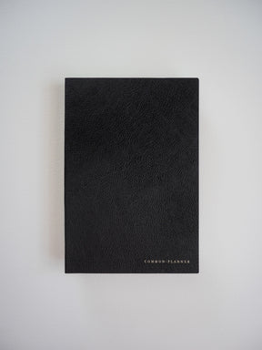 Minimalist matte black A5 planner with a subtly textured cover and the centered, debossed brand name "COMMON: PLANNER."