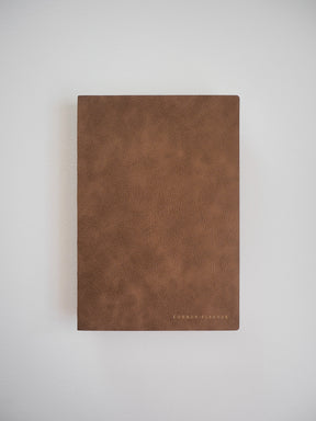 Elegant A5 vertical planner with a warm, cognac faux-leather cover, featuring minimalist branding centered near the bottom edge.