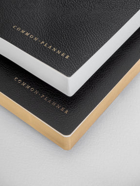 Two minimalist Common Planners stacked, showcasing textured black and dark gray covers with gold-edged bindings and subtle branded lettering.