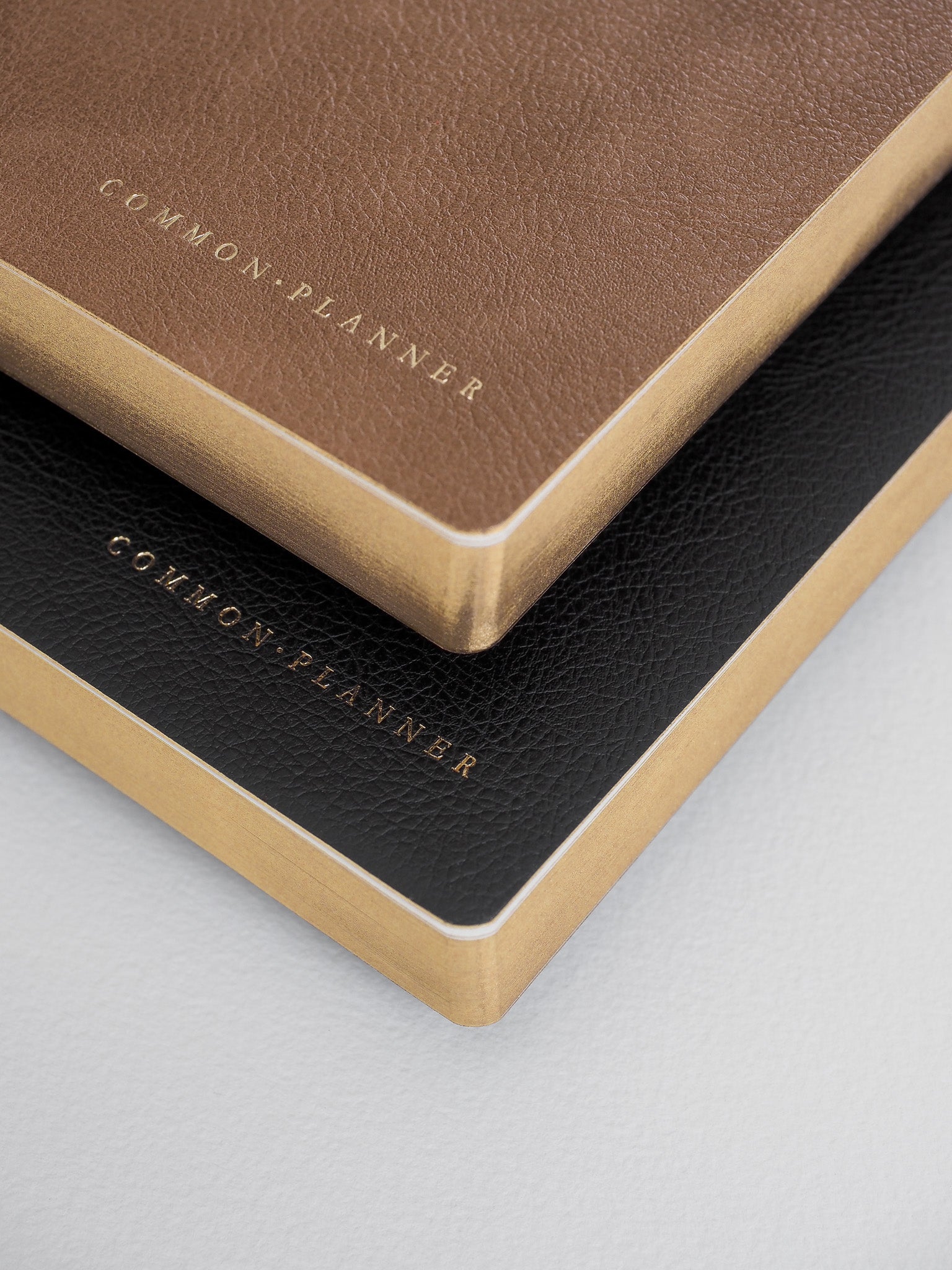 A set of two stacked A5 planners featuring matte brown and black covers, both accented with elegant gold foil lettering that reads "Common Planner."