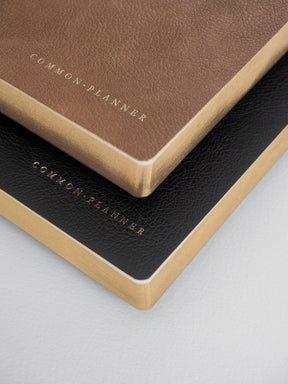 A set of two stacked A5 planners featuring matte brown and black covers, both accented with elegant gold foil lettering that reads "Common Planner."