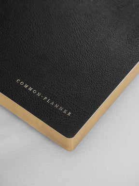 A sophisticated black planner with a matte cover, featuring elegant gold foil lettering that reads "COMMON • PLANNER" and accented with gold trim.