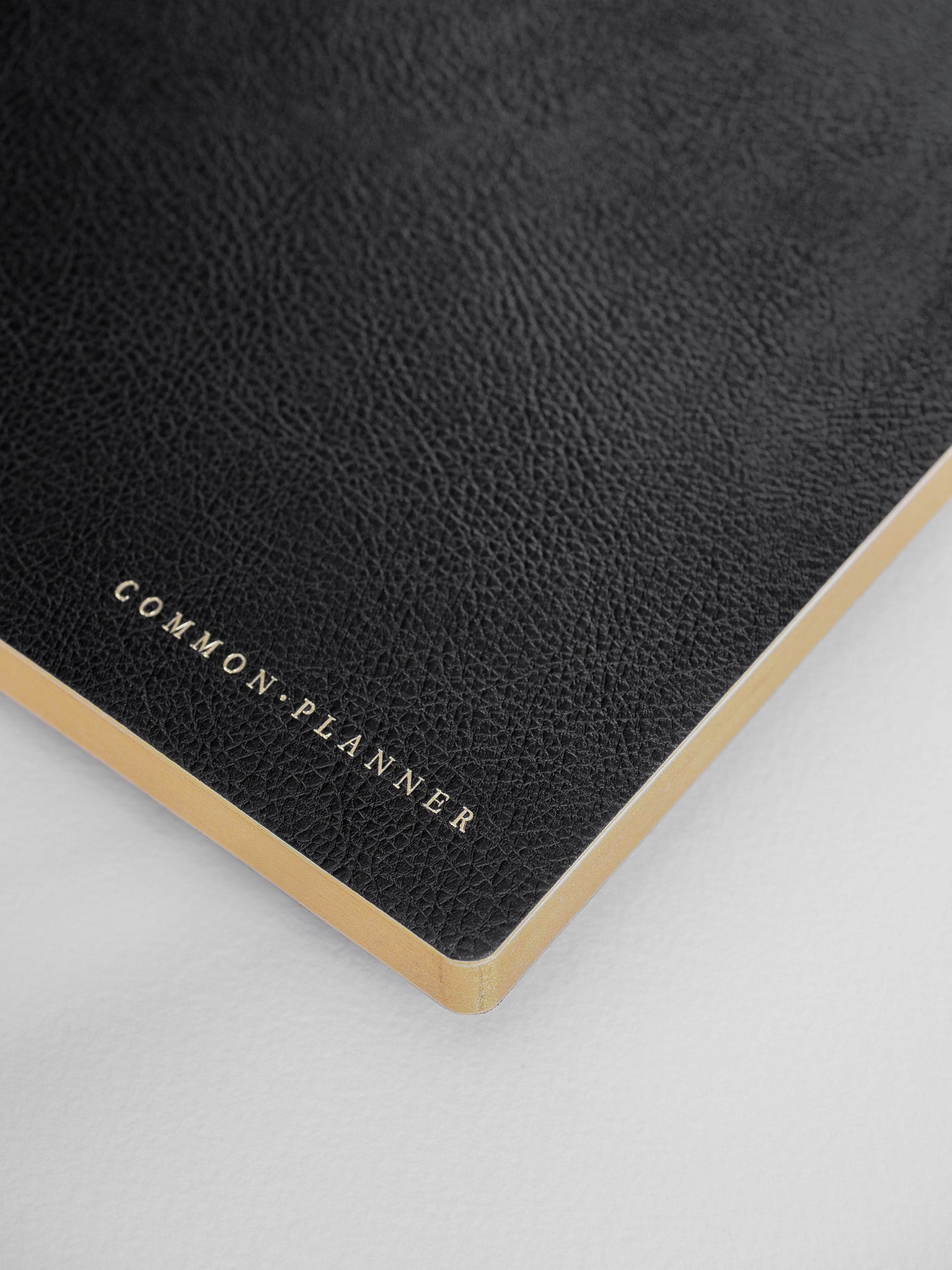Sleek, dark charcoal planner cover featuring elegant gold foil lettering that reads "COMMON PLANNER," accented by a natural tan binding edge.
