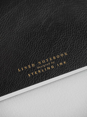 Black A5 notebook featuring a luxurious faux leather texture and elegant gold foil lettering that reads "LINED NOTEBOOK designed by STERLING INK."
