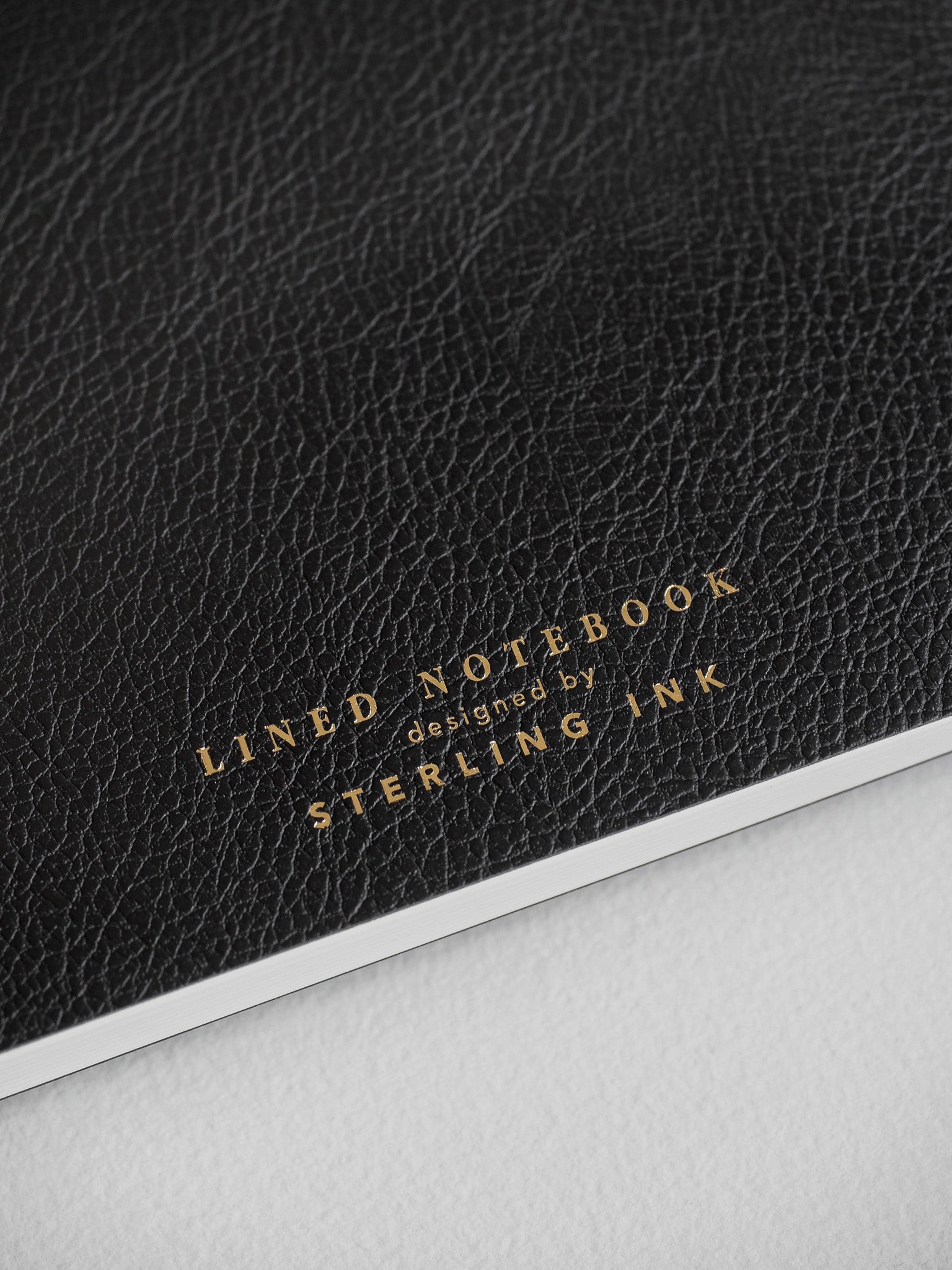Elegant black textured notebook featuring gold foil lettering that reads "LINED NOTEBOOK designed by STERLING INK."