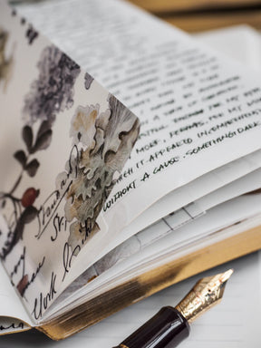A close-up of an open notebook or journal with handwritten text on the pages and a floral-patterned page folded over, showing botanical illustrations and cursive writing. A gold-finish fountain pen nib and dark barrel are visible in the foreground on a light surface.