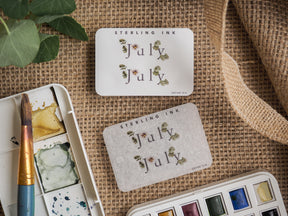 Decorative 'July' month sticker featuring the word in elegant script and delicate green botanical illustrations on a creamy off-white background.