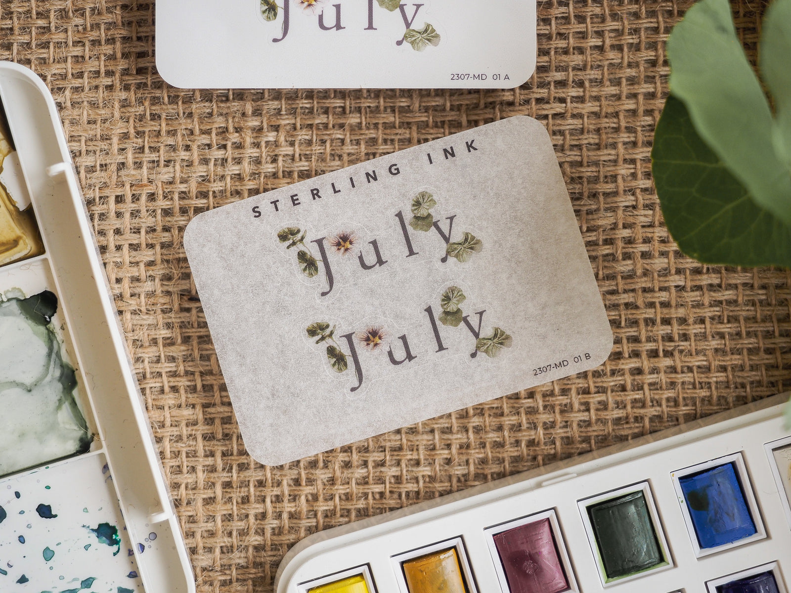 Cream-toned monthly sticker featuring "July" written in elegant script, adorned with delicate sage green botanical illustrations for journaling or planner use.