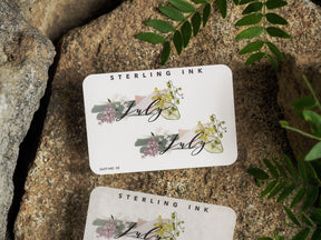 Watercolor floral sticker for July, featuring delicate pastel pink and green foliage surrounding the elegantly scripted month name.