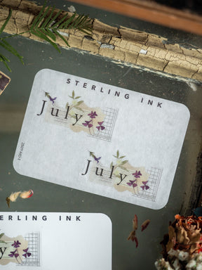 Adorable "July" month sticker perfect for planners, featuring the text surrounded by delicate purple wildflowers and natural green botanical accents.