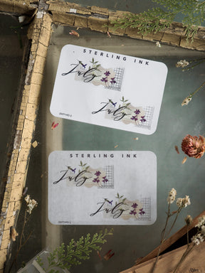 Elegant month sticker set for July, featuring the word "July" in sweeping cursive calligraphy adorned with purple floral accents and geometric designs.