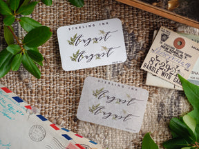 August month stickers featuring elegant script lettering, bordered by natural green watercolor foliage on various creamy white and rustic kraft paper backgrounds.