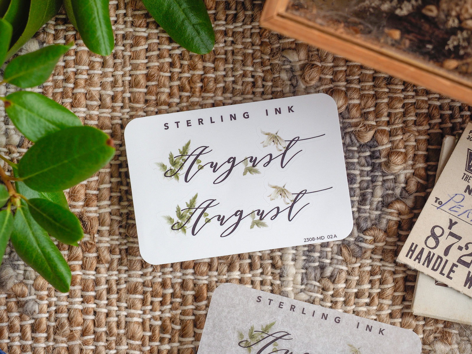 Decorative white month sticker featuring the word "August" in elegant, flowing script, accented by delicate watercolor green botanical leaf illustrations.