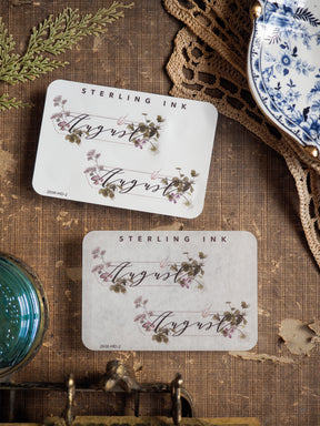 Elegant monthly sticker featuring "August" written in script font, surrounded by delicate watercolor illustrations of mauve and green botanicals.