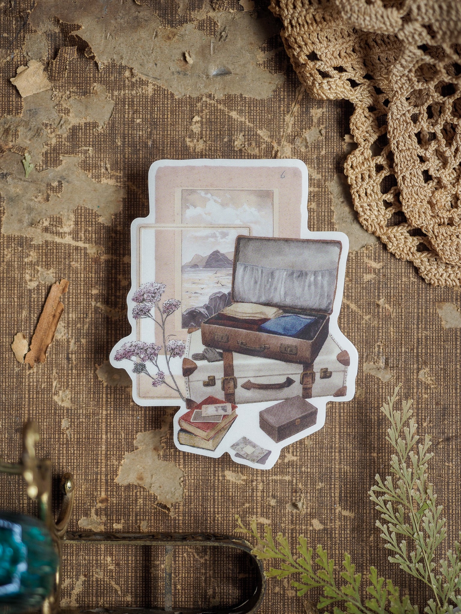Vintage die-cut sticker featuring an open, rustic suitcase set against a delicate background with stacked books and dried floral accents, perfect for travel journaling.