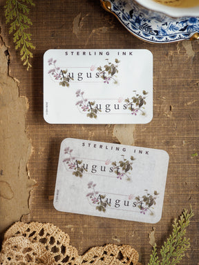 Three coordinating August month stickers featuring elegant botanical watercolor illustrations of purple blooms and green foliage on a soft cream background.