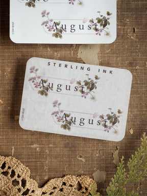 Delicate August month stickers with an elegant floral wreath design, featuring printed lavender and green wildflower illustrations on a pale cream background.