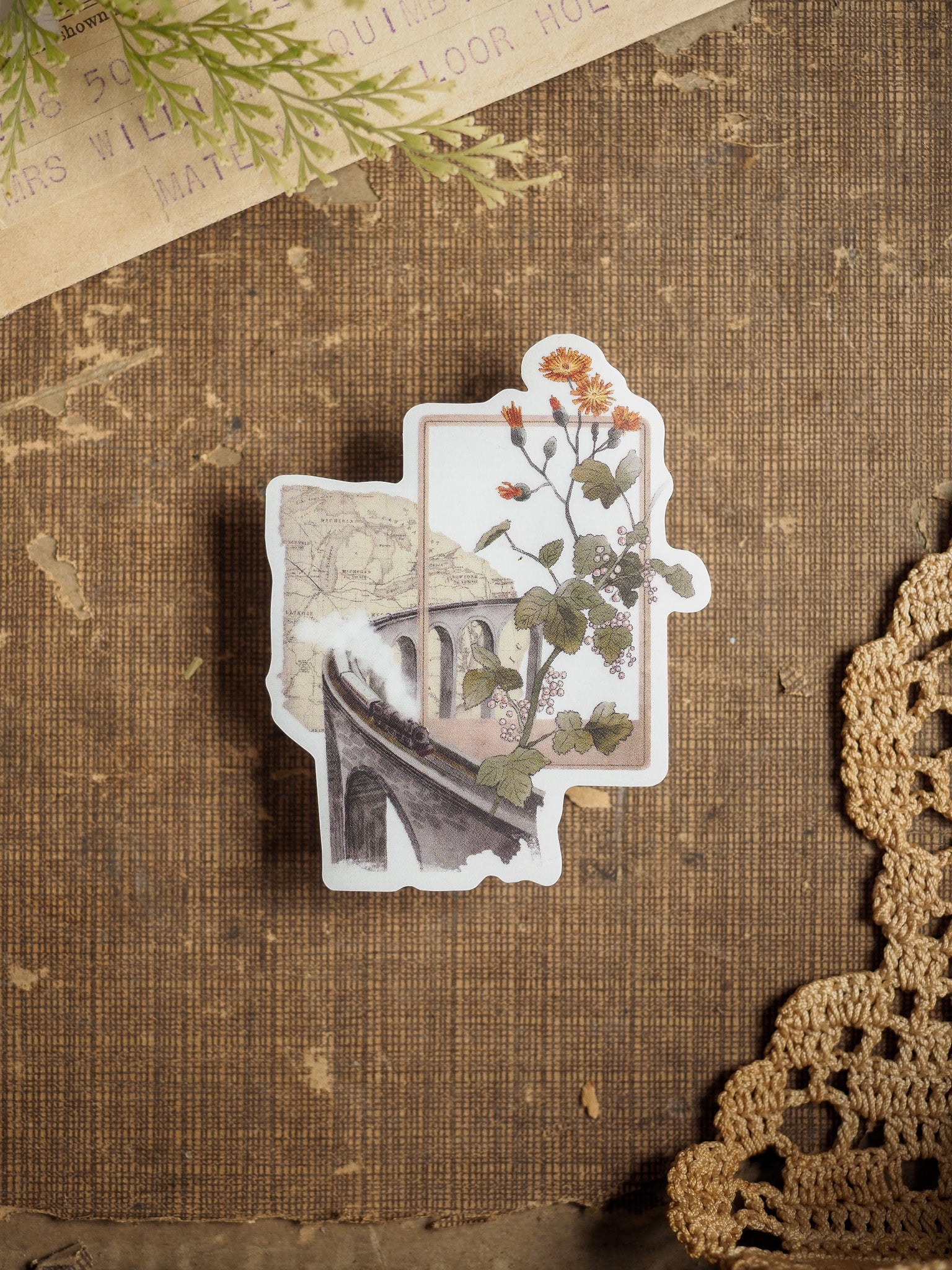Vintage die-cut vinyl sticker showing a scenic train crossing a rustic bridge, accented with delicate autumnal flowers and green foliage.