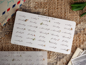 Set of decorative calendar stickers for August 2023, featuring "Monday" through "Sunday" written in elegant calligraphy and accented with delicate hand-drawn floral motifs.