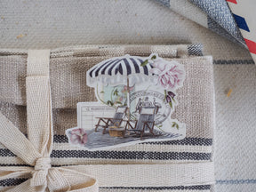 A sunny travel sticker featuring striped beach chairs, a striped umbrella, and delicate pink floral accents, perfect for decorating planners or scrapbooks.