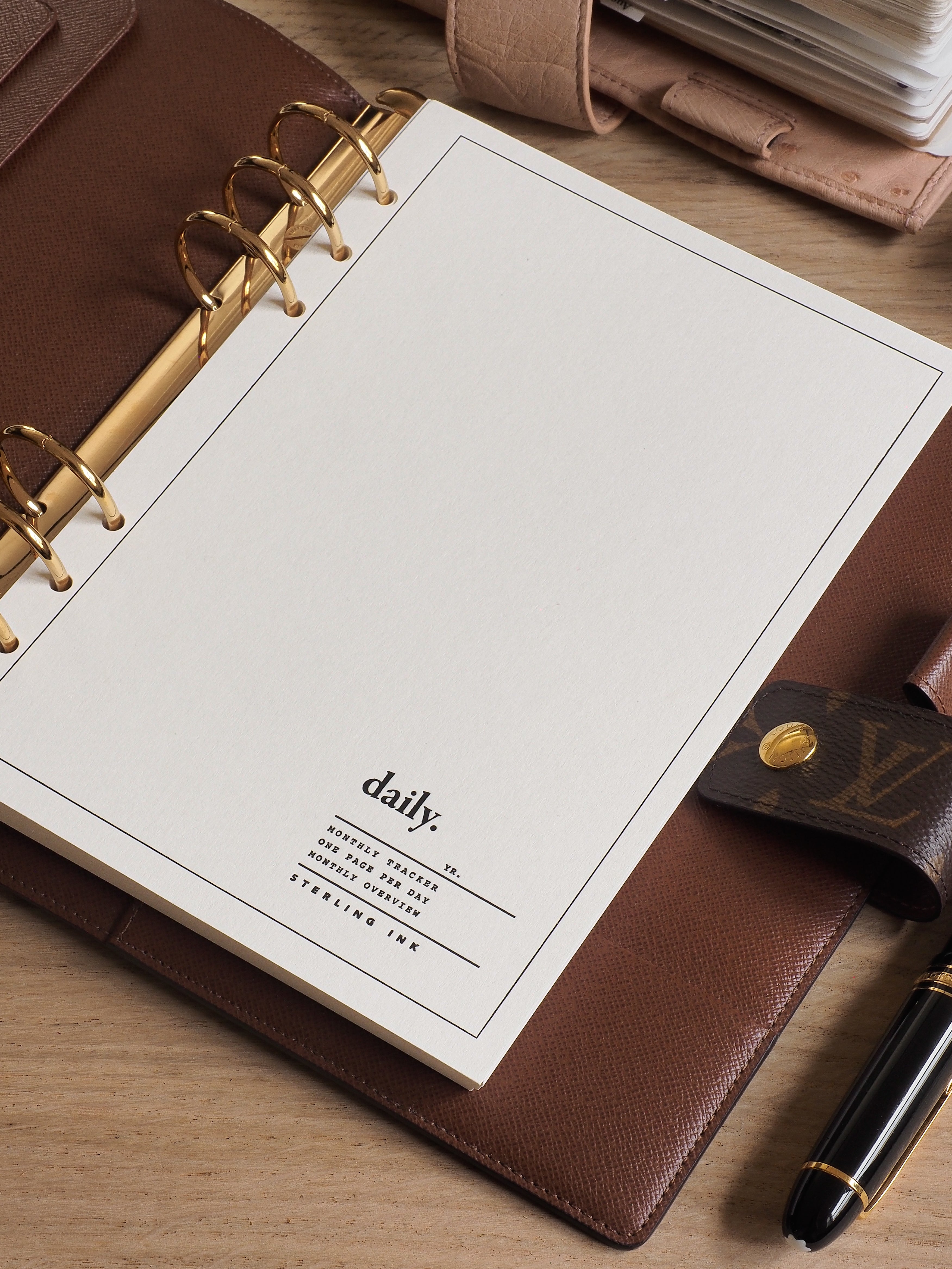 A brown leather-bound daily planner notebook with gold spiral binding, open to a cream page featuring the branded text "daily. MONTALLY TRACKER."
