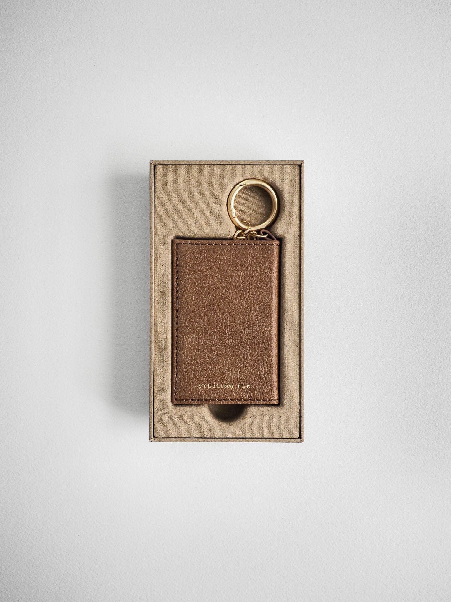 A brown leather card holder with a gold key ring, presented in its kraft paper packaging box against a light gray background.
#Color_Caramel