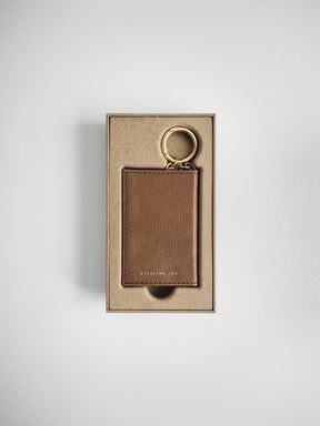 A brown leather card holder with a gold key ring, presented in its kraft paper packaging box against a light gray background.
#Color_Caramel
