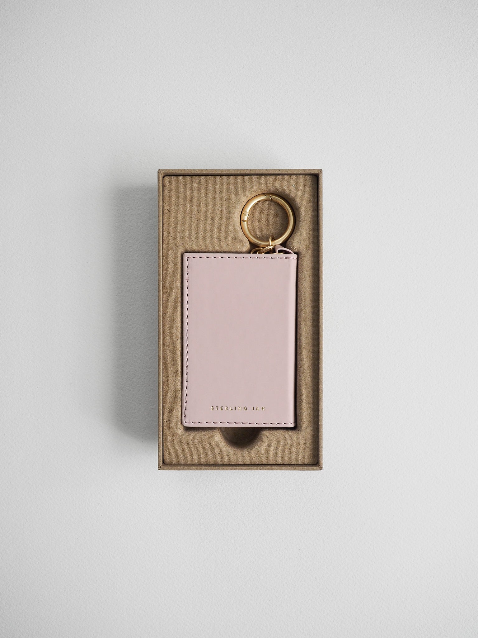 A light pink leather card holder keychain with a gold split ring, presented inside a brown kraft paper gift box on a light gray background.
#Color_Fairyfloss