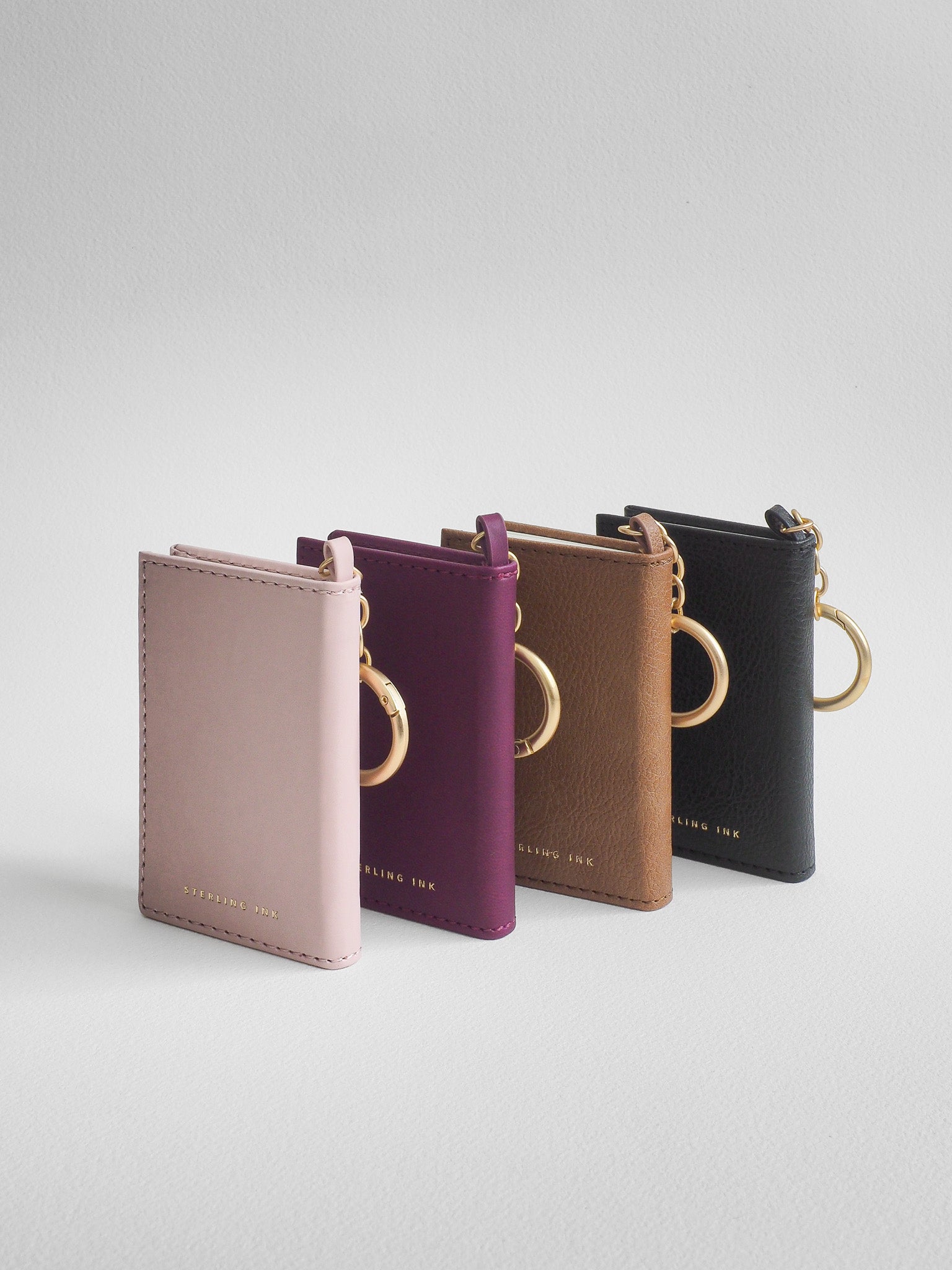 Four slim leather card holder/key case keychains in dusty pink, plum purple, tan brown, and black, arranged in a row on a light gray background. Each case features a gold-tone metal key ring and a small gold logo at the bottom.