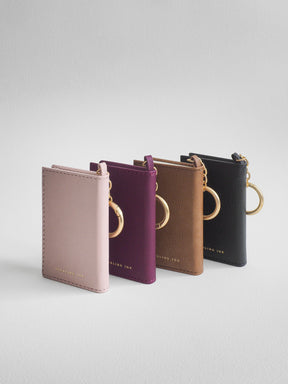 Four slim leather card holder/key case keychains in dusty pink, plum purple, tan brown, and black, arranged in a row on a light gray background. Each case features a gold-tone metal key ring and a small gold logo at the bottom.