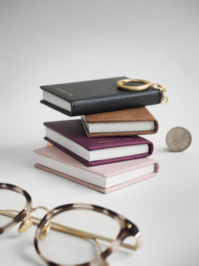 A stack of four mini faux-leather passport holder keychains in black, brown, purple, and pink, each designed to look like a book with a gold keyring on the spine. A pair of tortoiseshell eyeglasses and a coin are styled in the foreground on a light gray background.