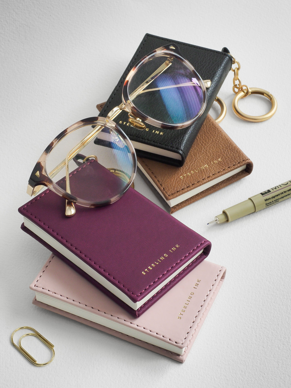 Four small leather-bound notebooks in black, brown, purple, and pale pink, arranged on a light gray surface with gold-tone glasses, a pen, paper clips, and a keychain charm. The notebooks show the brand name Sterling Ink.