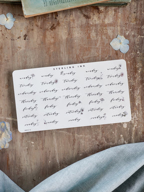 Decorative sheet of white planner stickers featuring black cursive typography for weekdays and delicate accompanying floral sprigs, ideal for journaling and organization.