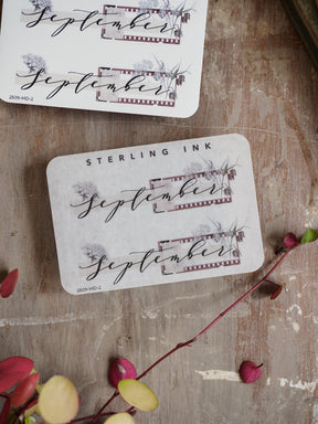 White planner stickers featuring "September" in graceful calligraphy, accented by a rustic, sepia-toned banner and delicate autumnal floral watercolor graphics.