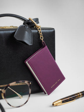 A purple leather passport holder keychain with gold hardware and a small gold logo, shown hanging from a black textured handbag with eyeglasses and a pen on a light gray background.
#Color_Mulberry