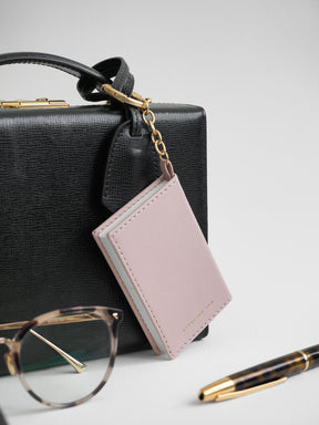 A light pink leather card holder wallet with gold-tone hardware, hanging from a black textured handbag. A pair of tortoiseshell eyeglasses and a gold-accented pen are styled on a light gray background.
#Color_Fairyfloss