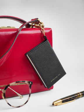 A red structured handbag with a black leather luggage tag attached by gold hardware, styled with tortoiseshell eyeglasses and a gold-and-brown pen on a light gray background.
#Color_Onyx